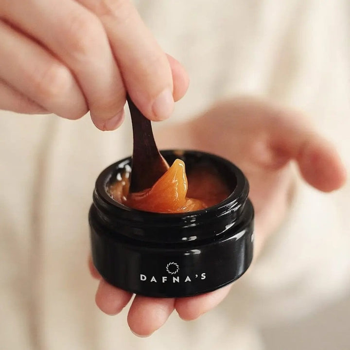 Open, black Dafna's Recovery Cleanser screw-top jar - one hand stirs the contents with a spatula. 