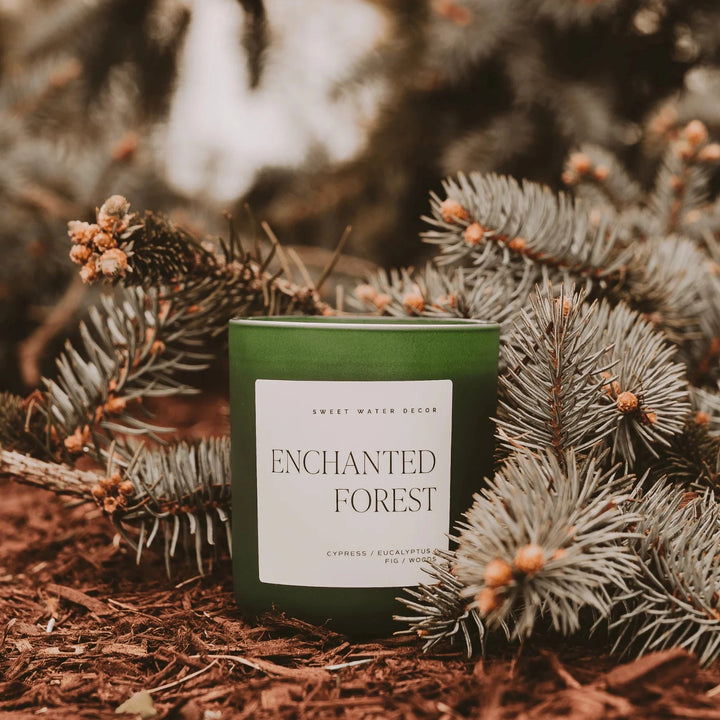 The "Enchanted Forest" candle from Sweet Water Decor in green frosted glass stands on a forest floor surrounded by small fir branches.