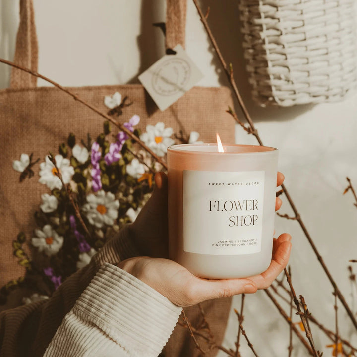 Candle in cream-colored milk glass "FLOWER SHOP" by Sweet Water Decor is balanced on a hand, behind it flowers, twigs and a jute bag.