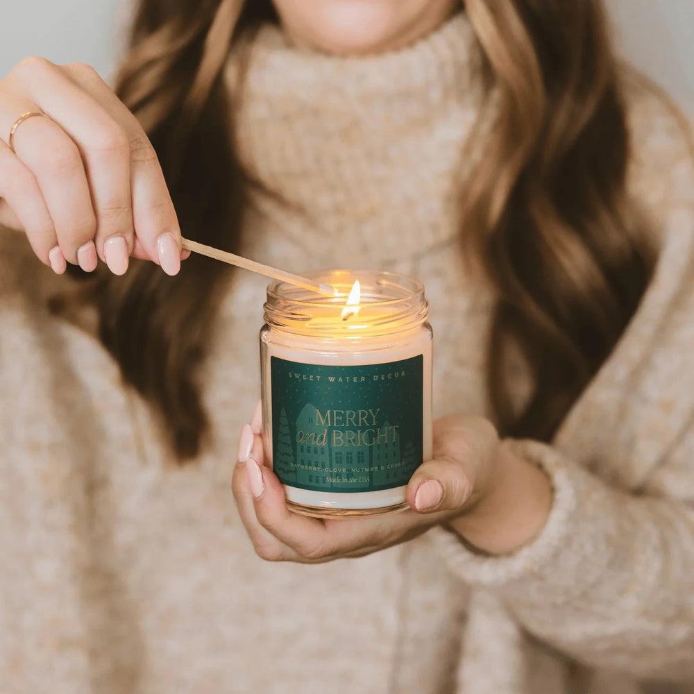 A "MERRY and BRIGHT" candle from Swet Water Decor is held by a woman and lit with a match.