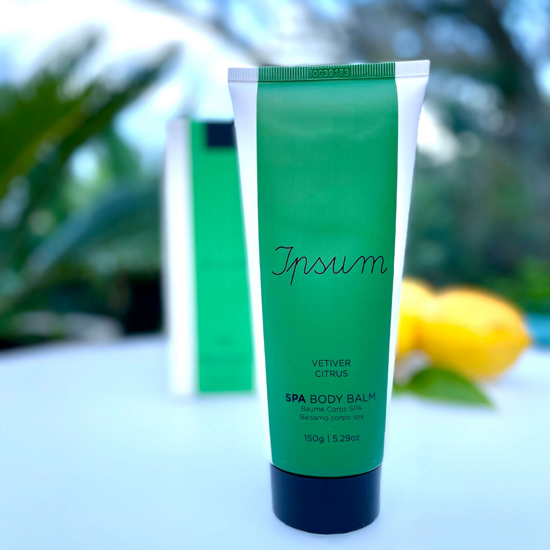 Green tube with blue lid of Ipsum Spa Body Balm stands on a white table, with blurry bushes and lemons in the background.