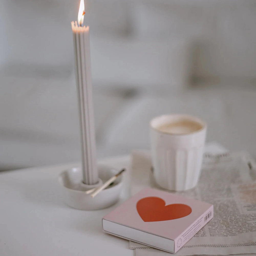 Streichholzschachtel mit Herz, liegend auf einer Zeitung, daneben eine brennende Stabkerze und ein Becher Kaffee.