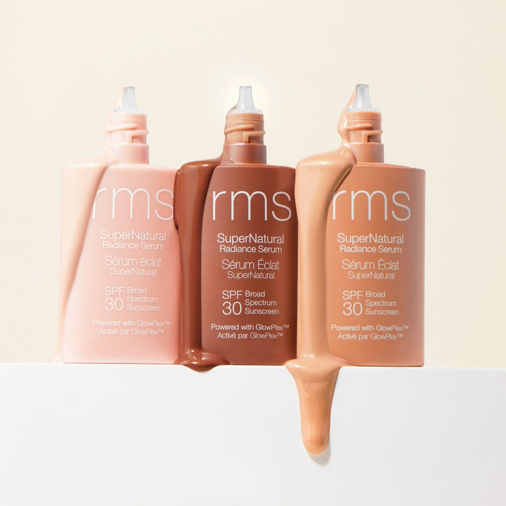 3 open bottles of RMS SuperNatural Radiance Serum with contents flowing out of them stand against a light background.