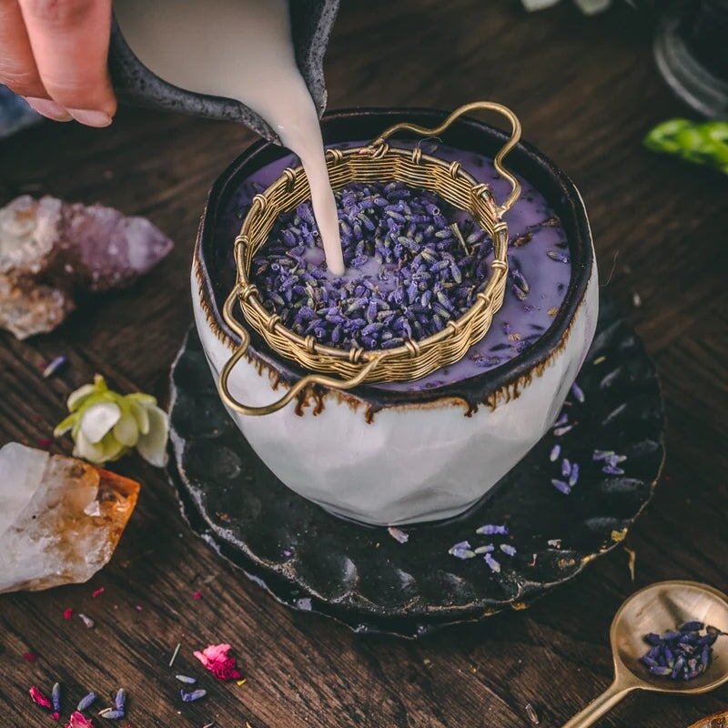 Anima Mundis Wooven Brass Large Tea Strainer - large tea strainer filled with lavender is infused with milky liquid in a large cup, stands on a wooden table, decorated with gemstones and flower petals.