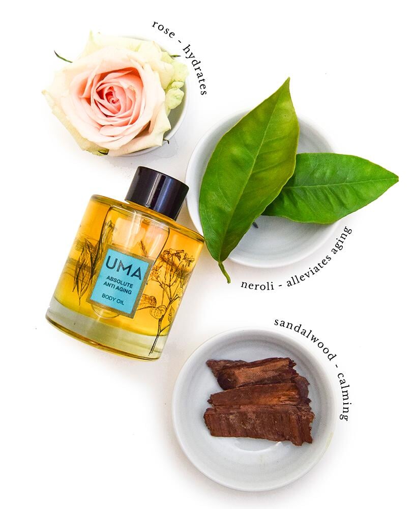 A bird's eye view of the UMA Absolute Anti Aging Body Oil bottle next to sandalwood, neroli leaves and a rose blossom.