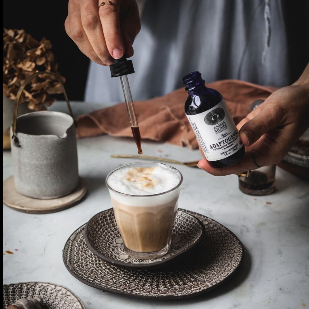 Eine Hand hält eine Anima Mundi Glasflasche Adaptogenic, die andere hält eine Pipette über ein Latte Macchiato Glas. North Glow