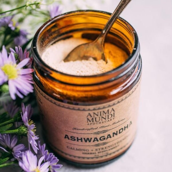 Offenes Anima Mundi Ashwagandha Schraubglas mit einem Löffel steht neben Blumen.