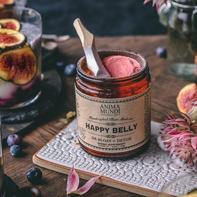 Offenes Belly Love Glas mit pinkem Pulver steht auf einem dekoriertem Holztisch.