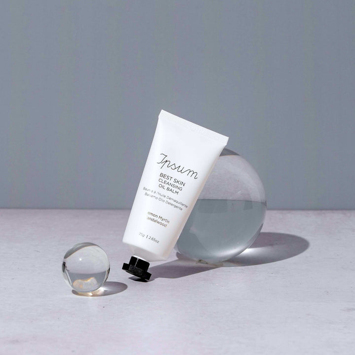 Closed tube of Ipsum Best Skin Cleansing Oil Balm leans against a glass sphere.