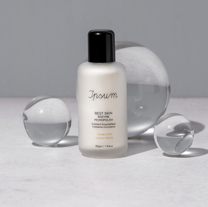 Ipsum Best Skin Enzyme MicroPolish frosted glass bottle with black lid stands between glass spheres of different sizes.