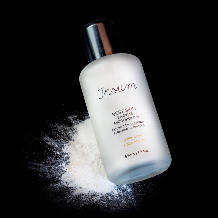 Ipsum Best Skin Enzyme MicroPolish frosted glass bottle with black lid with scattered powder against a black background.