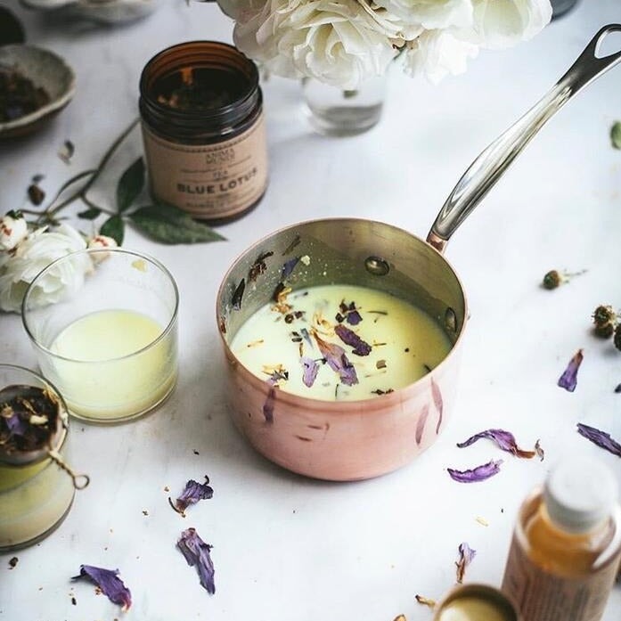 Copper stemmed pot of milky tea sprinkled with Blue Lotus tea leaves on white table next to a glass and open screw top jar of Anima Mundi Blue Lotus tea.  North Glow