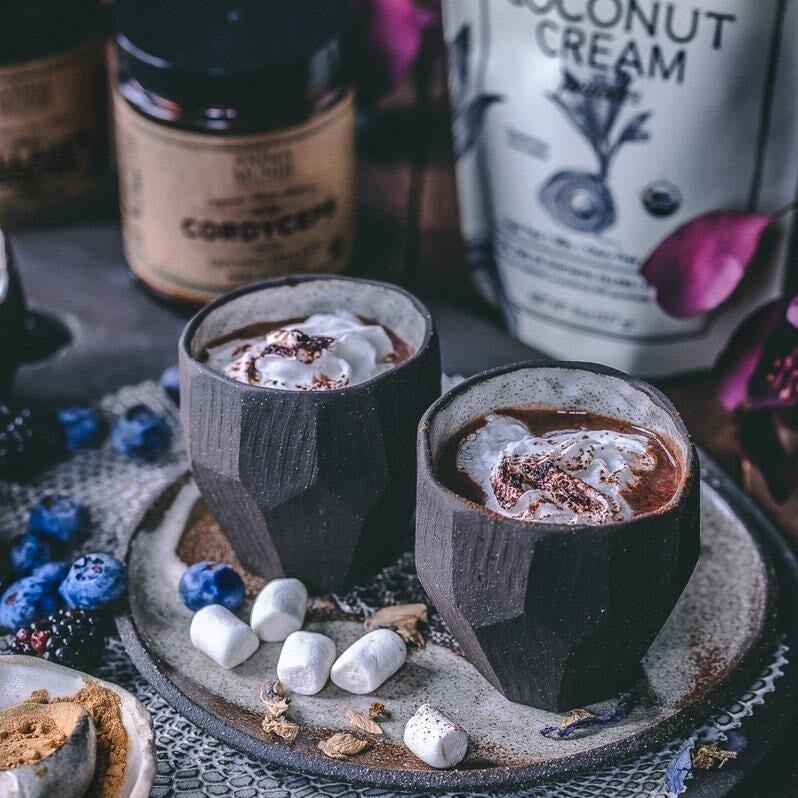 2 Steingutbecher mit cremigen Getränk stehen vor einer Anima Mundi Coconut Cream Powder Tüte.