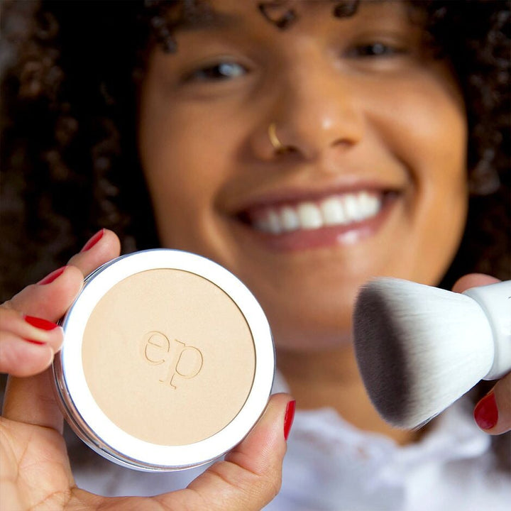 Blurred woman's face, in the foreground a hand holds an open can of Ere Perez Corn Setting Powder with a Kabuki brush.
