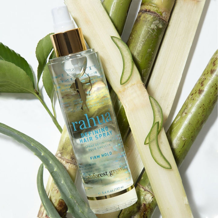 Rahua Defining Hair Spray bottle lies between bamboo stalks and aloe leaves against a white background.