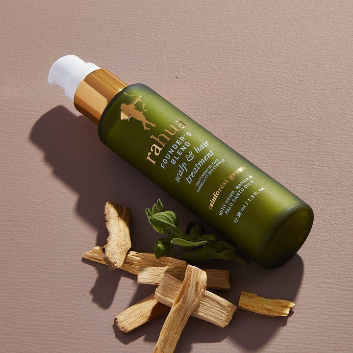 Founder`s Blend Scalp & Hair Treatment bottle lies next to wood chips and green tea leaves on a cream-colored background.