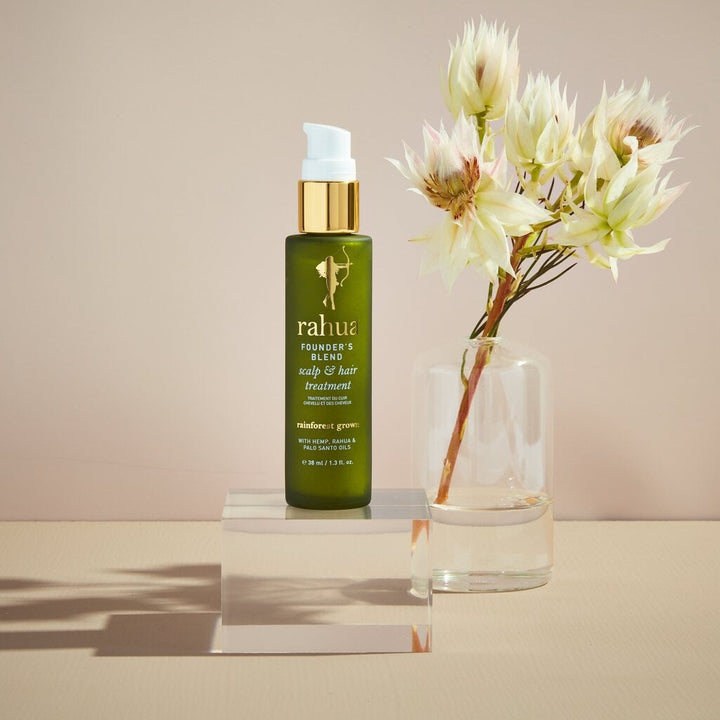 Founder`s Blend Scalp & Hair Treatment bottle stands next to a vase of white flowers against a cream-colored background.