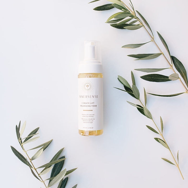 Between 3 olive tree branches lies a spray bottle of Innersense I Create Lift Volumizing Foam on a white background.