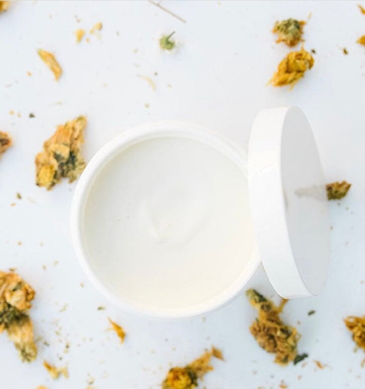 White, open jar filled with a cream from a bird's eye view, surrounded by dried petals.