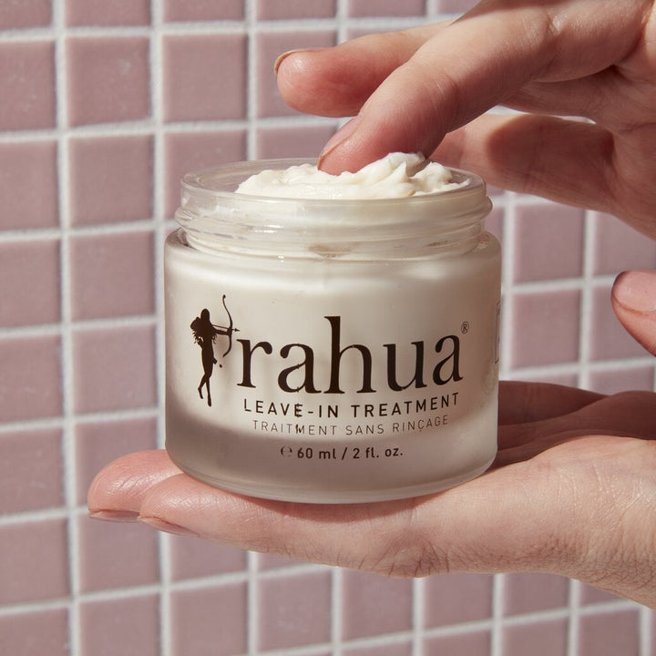 Rahua Leave-In Treatment jar, open, held by one hand, the other hand takes out product, in front of a pink tile background.