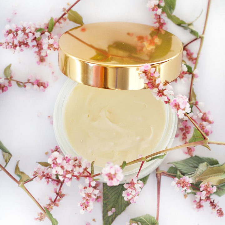 Offener Monsoon Paradise Body Soufflé Glastiegel mit angelehntem Deckel zwische Blüten auf weißem Untergrund.