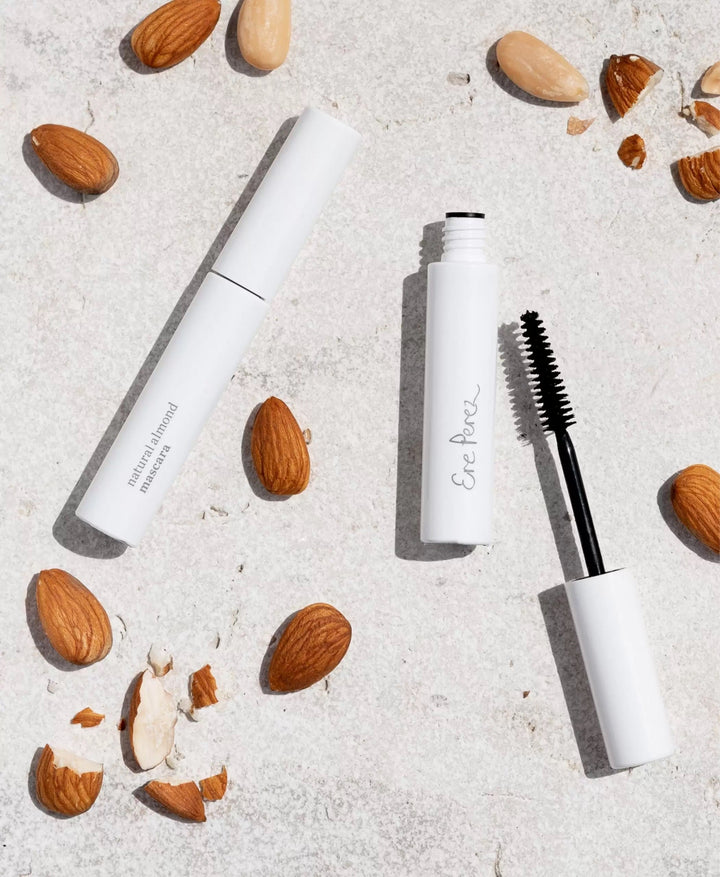 Eyelash brush and closed Ere Perez Natural Almond Mascara lies between almonds on a white base.
