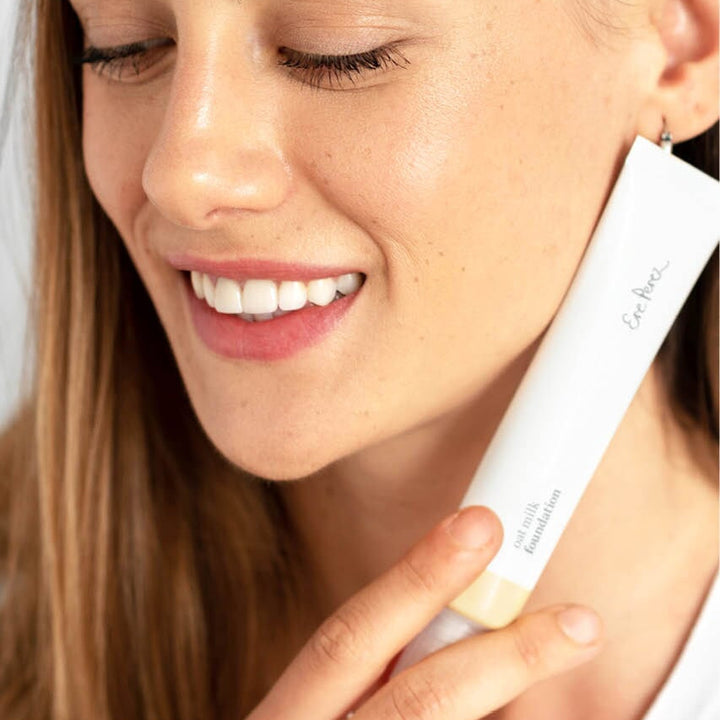 Close-up woman face, holding a tube of Oat Milk Foundation by Ere Perez.