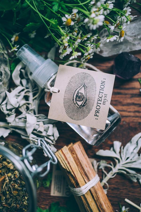 Anima Mundi Palo Santo Glasflasche mit einem großen Papierschild auf dem ein großes Auge abgebildet ist liegt auf verschiedenen Kräutern und Palo Santo Holzbündel.  North Glow