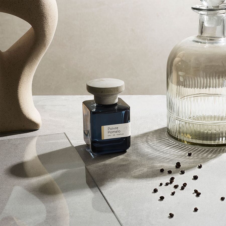 Dark blue glass bottle with gray lid "Poivre Pomelo" by Atelier Materi surrounded by a large glass jar, a glass plate on a gray background. North Glow