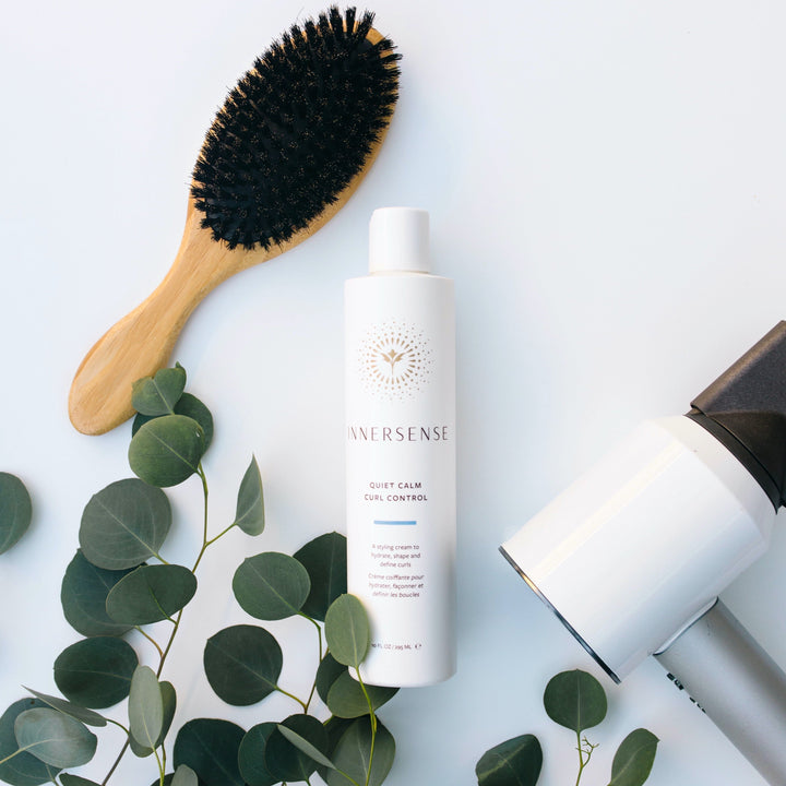 A bottle of Innersense Quiet Calm Curl Control surrounded by a hairbrush, hair dryer and a few twigs with green leaves on a white background.