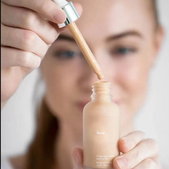 Glass bottle of the Ere Perez Quinoa Foundation in the foreground with a hand holding the pipette. Blurred female face in the background.