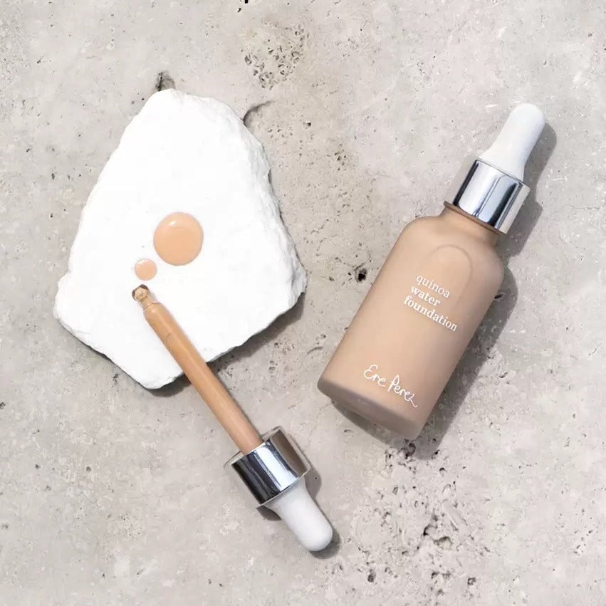 Glass bottle of Ere Perez Quinoa Foundation with pipette next to it and color and texture sample on a white stone.