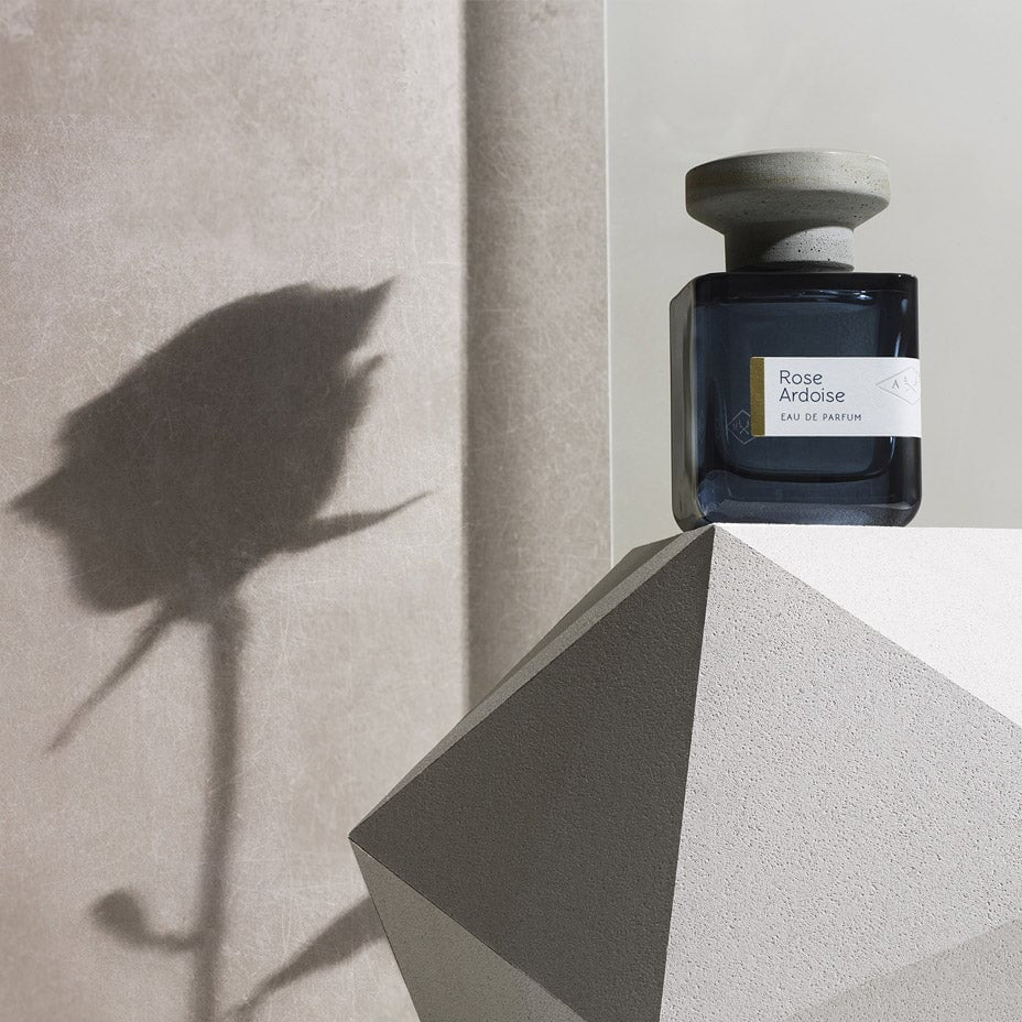 Dark blue glass bottle with gray lid "Rose Ardoise" by Atelier Materi with the shadow of a rose in the background. North Glow