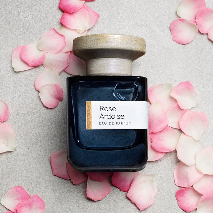 Dark blue glass bottle with gray lid "Rose Ardoise" by Atelier Materi, reclining, surrounded by rose petals.