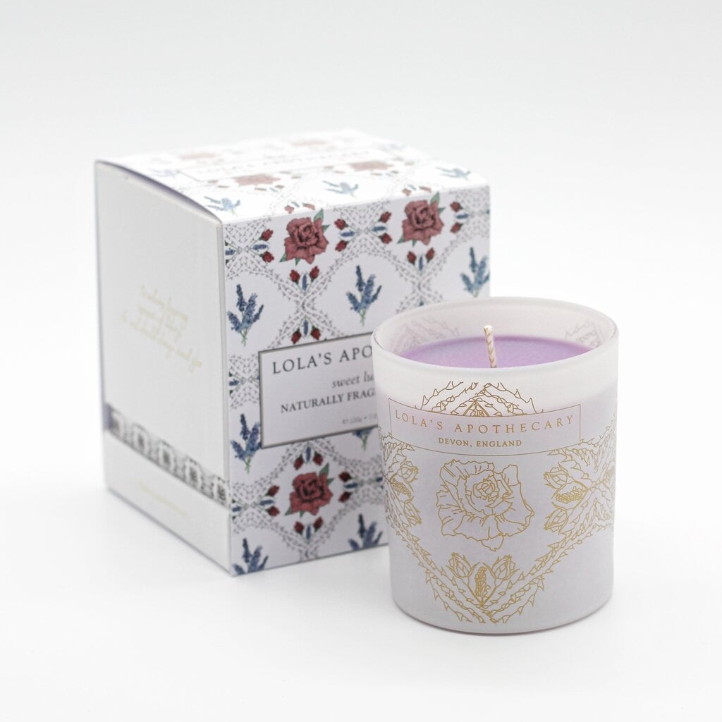 Lola's Apothecary Sweet Lullaby Duftkerze im verziertem Milchglas steht vor der Verpackung auf weißem Untergrund.