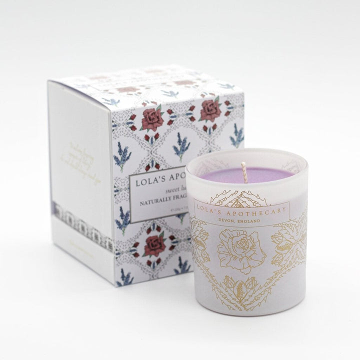 Lola's Apothecary Sweet Lullaby Duftkerze im verziertem Milchglas steht vor der Verpackung auf weißem Untergrund.