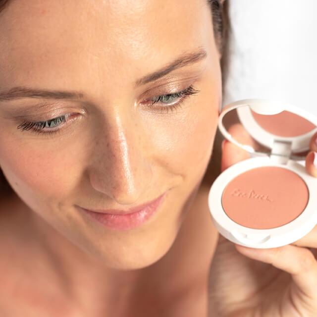 Close-up of a woman's face, she is holding an open can of Ere Perez Tapioca Cheek Color.