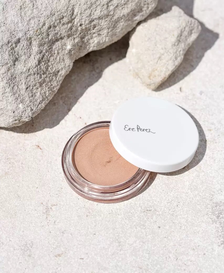 Open jar Ere Perez Vanilla Highlighter lies in the sand between stones.