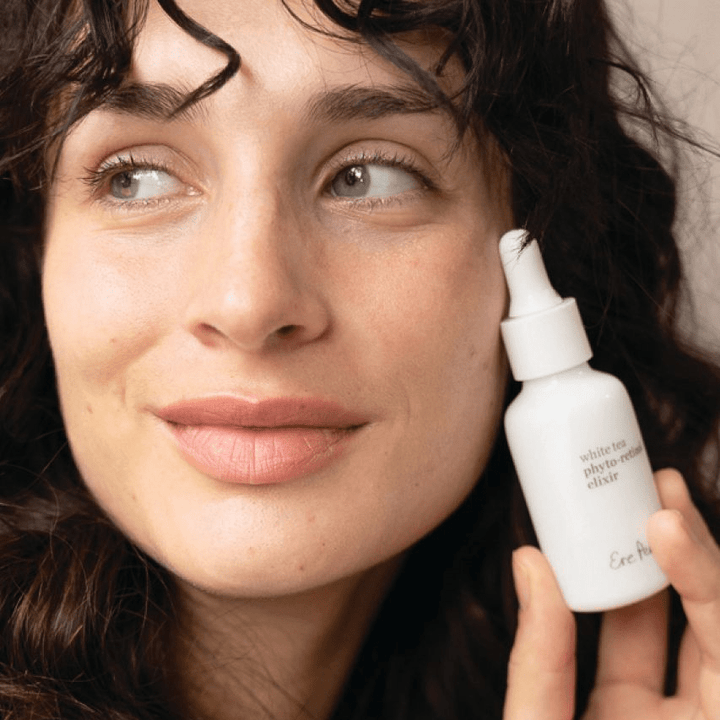 Close-up of a woman's face, holding a bottle of Ere Perez White Tea Phyto-Retinol Elixir.