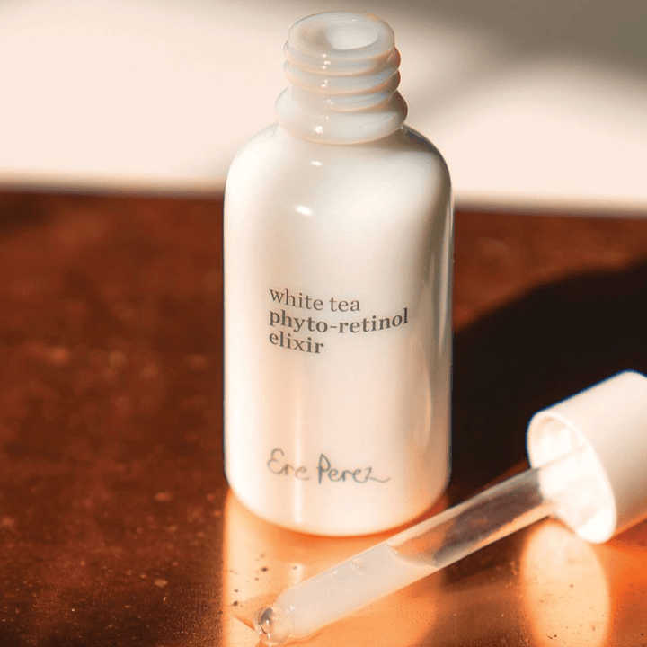 Open bottle of Ere Perez White Tea Phyto-Retinol Elixir with pipette lies on a bronze-colored background.